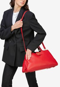 Rød læder håndtaske med en struktureret form, guld hardware accenter, og en aftagelig skulderrem, holdt op mod en sort blazer.