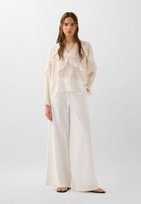 Blouse blanche légère avec des couches volantes et de petits pois dorés, associée à un pantalon blanc ample. Le mannequin se tient devant un fond uni.