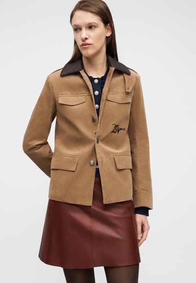 Γυναίκα που φοράει μια μπεζ ζακέτα από κορντόρι με τσέπες με καπάκι και σκούρο γιακά, συνδυασμένη με μια βουργουνδί δερμάτινη φούστα και μαύρες καλσόν.