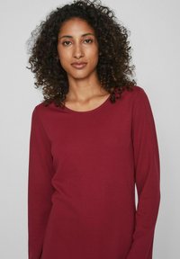 Top de manga larga en burdeos de tela suave, con escote redondo, textura lisa y corte ligeramente holgado. Sin patrones visibles ni herrajes.