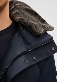 Marineblauwe jas met een pluche grijze bontvoering in de capuchon, voorzien van een gladde buitenstof, ritssluiting en metalen hardwareaccenten.