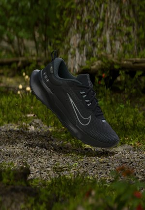 Zapatilla deportiva negra con un swoosh reflectante flotando sobre grava y vegetación en un entorno forestal al aire libre.
