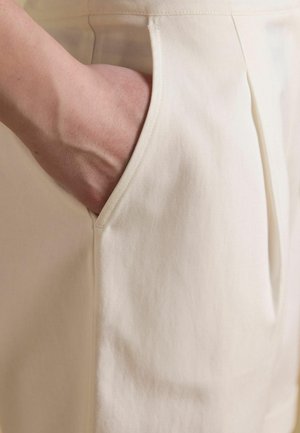 Hand teilweise in die Vordertasche beiger oder off-weißfarbener Stoffhose mit sichtbaren Nähten gesteckt.