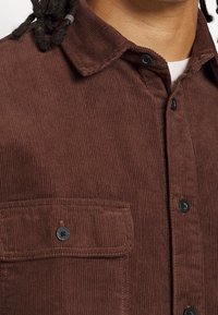 Braunes Cordhemd mit einem Button-Down-Kragen, schwarzen Knöpfen und einer Brusttasche mit Knopfverschluss. Strukturierter Stoff mit vertikalen Streifen.