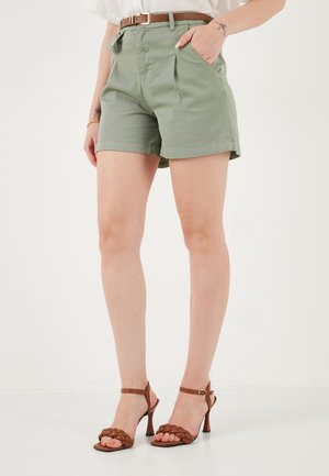 Vrouw die olijfgroene shorts met hoge taille en riem draagt, gecombineerd met bruine sandalen met gevlochten bandje en hoge hak, staand tegen een witte achtergrond.