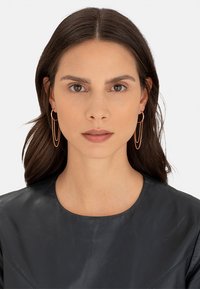 Boucles d'oreilles créoles en or rose avec un détail de chaîne cascade, portées par un modèle en haut en cuir noir. Le design présente des finitions lisses et des lignes simples.