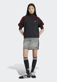 Chaqueta atlética negra con rayas rojas, falda de mezclilla gris, calcetas negras hasta la rodilla con rayas blancas y zapatillas negras/blancas.