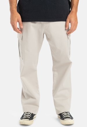 Beige cargo byxor med flera sidofickor, tillverkade av bomull, med rak passform och en jämn textur. Bärs med svarta sneakers.