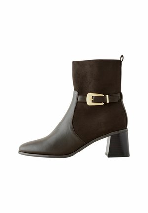 Brązowy skórzany i zamszowy botek za kostkę z ostrym noskiem, słupkiem i dekoracyjnym paskiem z klamrą wokół kostki.