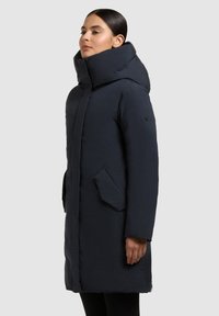 Abrigo acolchado azul marino hecho de nylon, con cuello alto, cierre de cremallera, bolsillos laterales y un corte holgado. Textura suave con acabado mate.