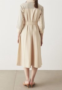 Abito midi beige con vita regolabile e pannelli testurizzati sulle spalle. Realizzato in un tessuto leggero, presenta una silhouette fluida.