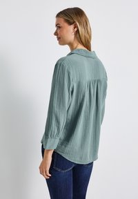 Hellgrüne, strukturierte Bluse mit Kragen, Dreiviertelärmeln und geraffter Rückennaht, kombiniert mit dunkelblauen Jeans.