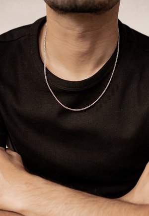 Silberne Kettenhalskette, die auf einem nackten Hals ruht, mit einem dünnen, gedrehten Design. Die Trägerin trägt ein geripptes schwarzes Shirt und hat die Arme verschränkt.