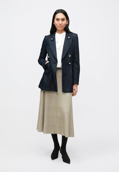 Σκούρο τζιν σακάκι διπλής σειράς με ασημένια κουμπιά, φορεμένο πάνω από λευκό πουκάμισο και πλισέ μπεζ φούστα. Μαύρες καλσόν και μυτερές γόβες ολοκληρώνουν το σύνολο.