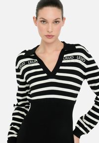 Robe polo en tricot rayée noir et blanc avec un col en V, ornée de détails de logo et d'accents en perles sur les poignets. Design ajusté.