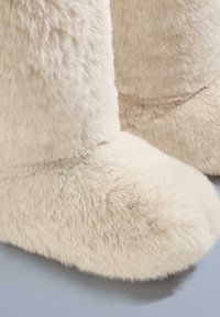 Krém színű, puha plüss csizma, puha, bolyhos textúrával és lekerekített orral. Az anyag vastagnak és kényelmesnek tűnik, beltéri használatra alkalmas.