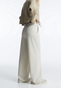 Beige cropped jasje gecombineerd met wijde, lichtbeige broek. De broek heeft achterzakken en een gladde, textuurachtige afwerking.