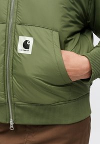 Zelena podložena jakna z rebrastim robom in manšetami; ima logotip na žepu na prsih in sprednji zadrgo.