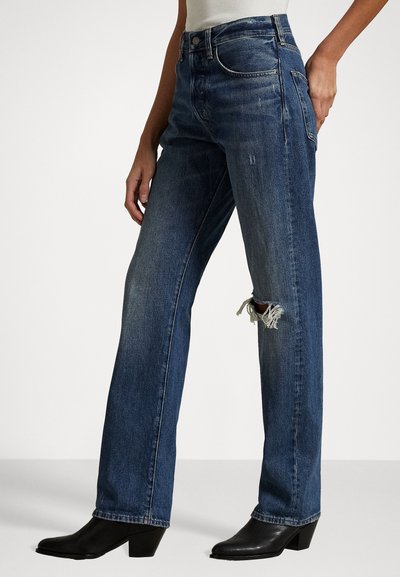 Blå denimjeans med løs passform, høy midje, et revnet detalj på kneet, og subtil falming. Brukt sammen med svarte hælboots.