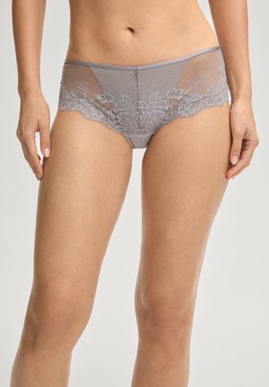 Vrouw draagt grijze kanten mesh hipsteronderbroek met bloemrijke borduursels en een klein sierseltje aan de voorkant, staand met ontspannen armen langs het lichaam.