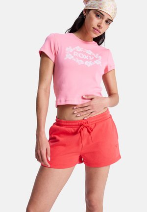 Młoda kobieta w różowej kwiecistej opasce na głowę, różowym skróconym t-shircie Roxy oraz czerwonych szortach z troczkami na białym tle.