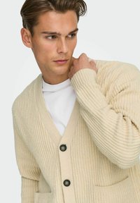 Beige gerippter Strickcardigan mit V-Ausschnitt, zwei vorderen Taschen und schwarzen Knöpfen. Über einem weißen T-Shirt getragen. Glatte Textur, lockere Passform.