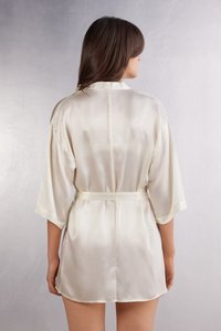 Satin kort kappa i elfenben, med skalkrage, vida ärmar och en knytbelte i midjan, med en mjuk, glänsande yta.