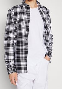 Zwart-wit geruit overhemd met lange mouwen, knoopsluiting en kraag. Draagt het over een effen wit t-shirt, gecombineerd met lichtgekleurde broek.