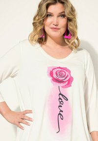 Top bianco a maniche lunghe con una grafica di una rosa rosa e la parola "amore" in script nero. Il tessuto ha una texture liscia.
