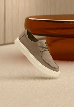 Zapato casual de ante marrón claro con cordones y suela blanca flotando sobre una alfombra beige y una mesa de madera marrón de fondo.