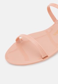 Sandale de couleur pêche en matériau plastique lisse et brillant. Présente deux fines lanières et une semelle ergonomique avec des détails cousus.
