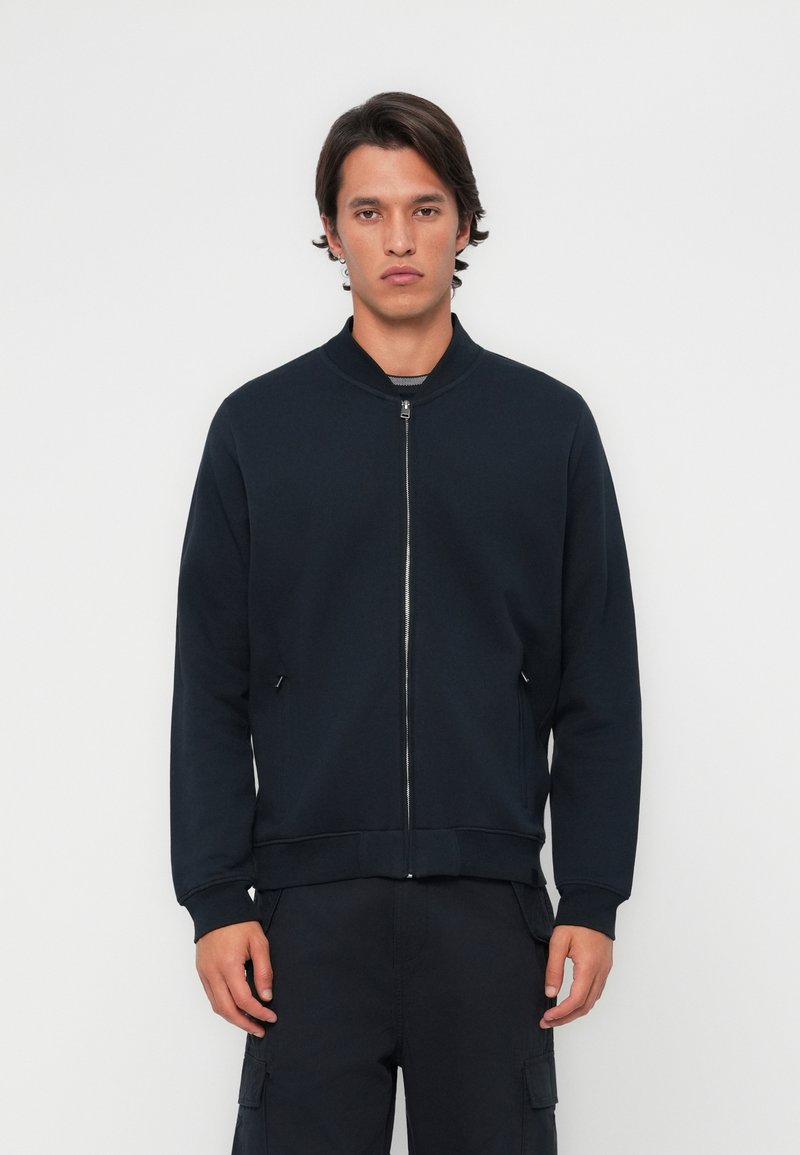Veste bomber zippée bleu marine avec col rond, dotée de poches latérales et de poignets côtelés, fabriquée en tissu lisse et doux.