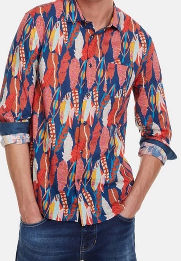 Homme portant une chemise à manches longues avec un motif de plumes colorées en rouge, bleu, blanc et jaune, manches retroussées, les mains dans les poches de son jean.