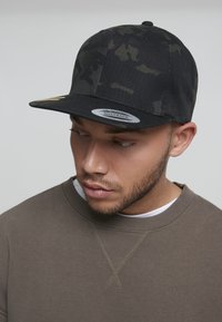 Fekete snapback sapka álcázott mintával, lapos szélllel és lélegző anyagból készült. Szürke pulóverrel viselve.