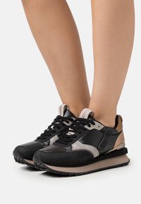 Zapatillas deportivas negras y grises con un diseño texturizado, materiales de cuero y ante, y una suela y un acabado en el talón de color beige en contraste.