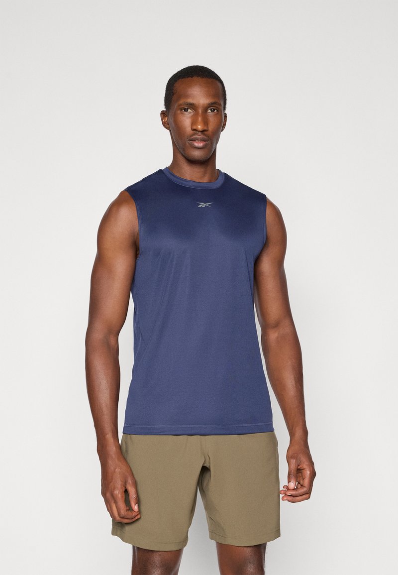 Débardeur de sport sans manches bleu marine en tissu léger, avec un col rond et un logo argenté sur la poitrine. Porté avec un short beige.