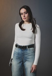 Weißes langärmeliges Oberteil mit Schulterdetails, kombiniert mit hellblauen High-Waist-Jeans und einem schwarzen Gürtel mit einer runden Schnalle. Schwarze Handschuh an der linken Hand.