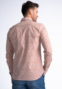 Camisa marrón con estampado, de cuello abotonado, mangas largas y un sutil diseño floral. Presenta un dobladillo redondeado y detalles con botones en los puños.