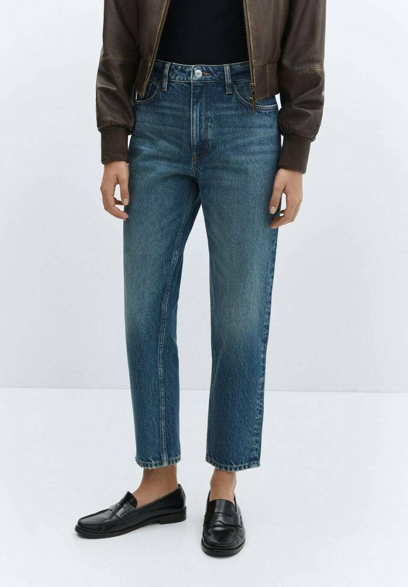 Eine Person in abgeschnittenen blauen Jeans, schwarzen Slippers, einem schwarzen Oberteil und einer braunen Lederjacke steht vor einem schlichten Hintergrund.