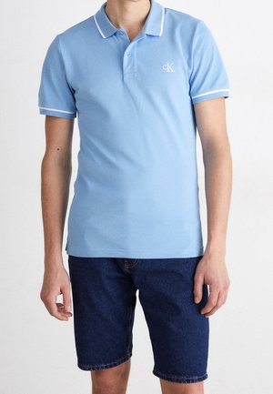 Homme portant un polo bleu clair à manches courtes avec bordure blanche et un short en jean bleu foncé, debout sur fond blanc.