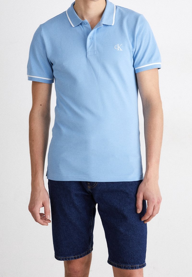 Homme portant un polo bleu clair à manches courtes avec bordure blanche et un short en jean bleu foncé, debout sur fond blanc.