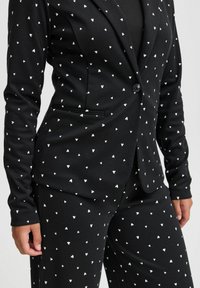 Svart blazer och byxset med små vita hjärtan, bärs med en svart topp under, vilket visar midjan och händerna.