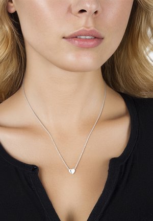 Primer plano de una mujer que lleva un collar con un colgante plateado en forma de corazón sobre una cadena fina, combinado con una blusa negra con un pequeño detalle de botón.