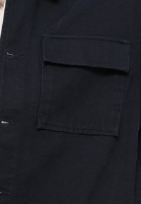 Σκούρος ύφασμα με μεγάλη τετράγωνη τσέπη και εμφανή κουμπότρυπες στην πλευρά, που δείχνουν λεπτομερή ραφή και υφή.