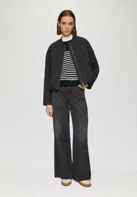 Schwarze Bomberjacke mit Reißverschluss, getragen über einem gestreiften Pullover, kombiniert mit dunklen, weit geschnittenen Jeans und beigen Schuhen. Glatte Stofftextur.