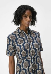 Camisa de manga corta con fondo azul y un gran estampado floral en marrón y crema. Presenta un cuello y un cierre frontal con botones.