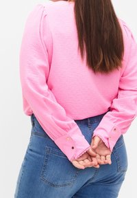 Chemise rose texturée à manches longues avec épaules bouffantes, ornée d'accents en boutons dorés et de bordure en dentelle aux poignets, associée à un jean bleu.