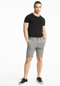 Pantaloncini grigi con risvolto e tasche laterali, abbinati a una t-shirt nera aderente e sneakers nere, in piedi su uno sfondo bianco.