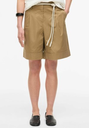 Osoba ubrana w beżowe szorty do kolan z białym paskiem ze ściągaczem, białą koszulę, czarne buty wsuwane oraz czarną opaskę na rękę.