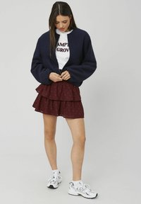 Veste bomber courte bleu marine, t-shirt blanc à motif graphique, jupe froncée bordeaux à plusieurs couches, et chaussures de sport blanches, créant une tenue décontractée.